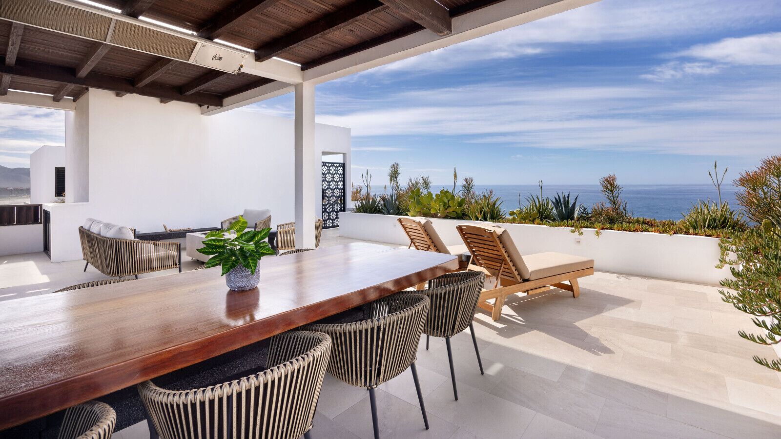 A modern outdoor dining area with a wooden table and chairs, a potted plant, and a view of the sea.