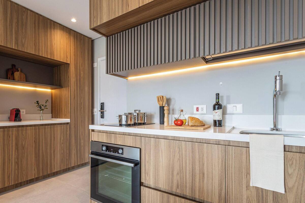Een moderne keuken met houten kastjes, een inbouwoven, een aanrecht met een wijnfles en een snijplank met groenten.