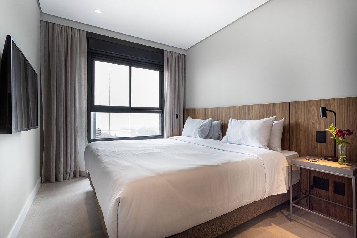 Een moderne hotelkamer met een groot bed, een aan de muur bevestigde televisie, een nachtkastje met lamp en een raam met gordijnen.