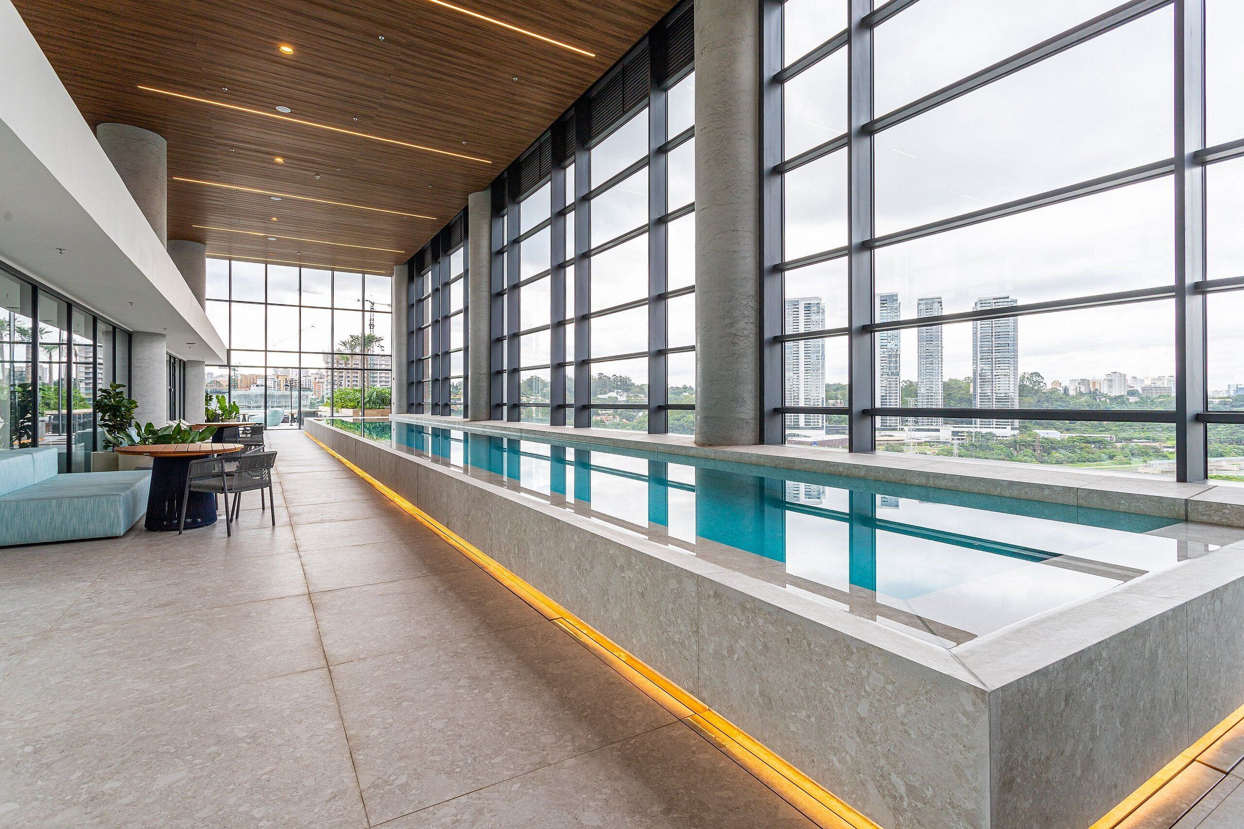 A modern indoor pool area with large windows, a long bench, and a seating area with tables.