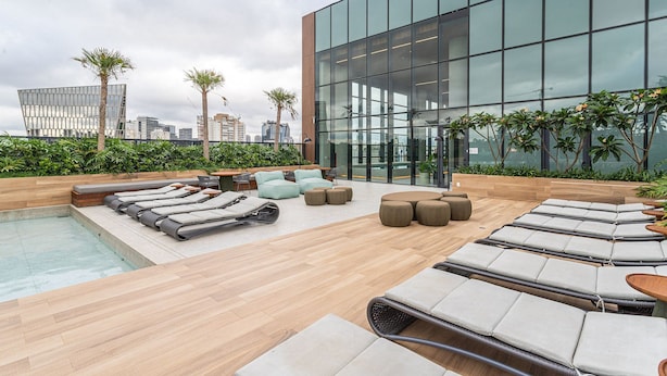A rooftop lounge with sun loungers, a pool, and a modern building in the background. - AV.NU by Viva - Residencial Alto Padrão próximo a Business Center na Nações Unidas. (São Paulo)