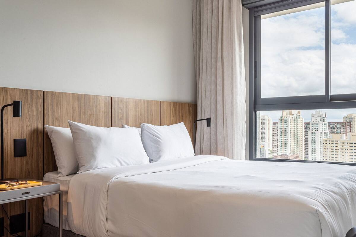 Een hotelkamer met een bed, een nachtkastje, een lamp en een raam met uitzicht op de stad.