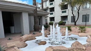 A courtyard with a water fountain, surrounded by rocks and desert plants, in front of a building with balconies and a bench. - Resort living in Scottsdale w heated pool #124 (Scottsdale)