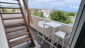 Ein Balkon mit einer Holztreppe, zwei weißen Stühlen und Blick auf das Meer und die Gebäude.