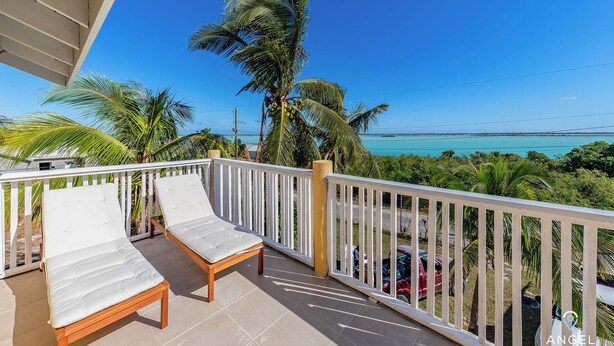 A balcony with two white cushioned chairs and a wooden table, overlooking a turquoise sea and palm trees. - Bright & Roomie Bottle Creek Apts (Bottle Creek)