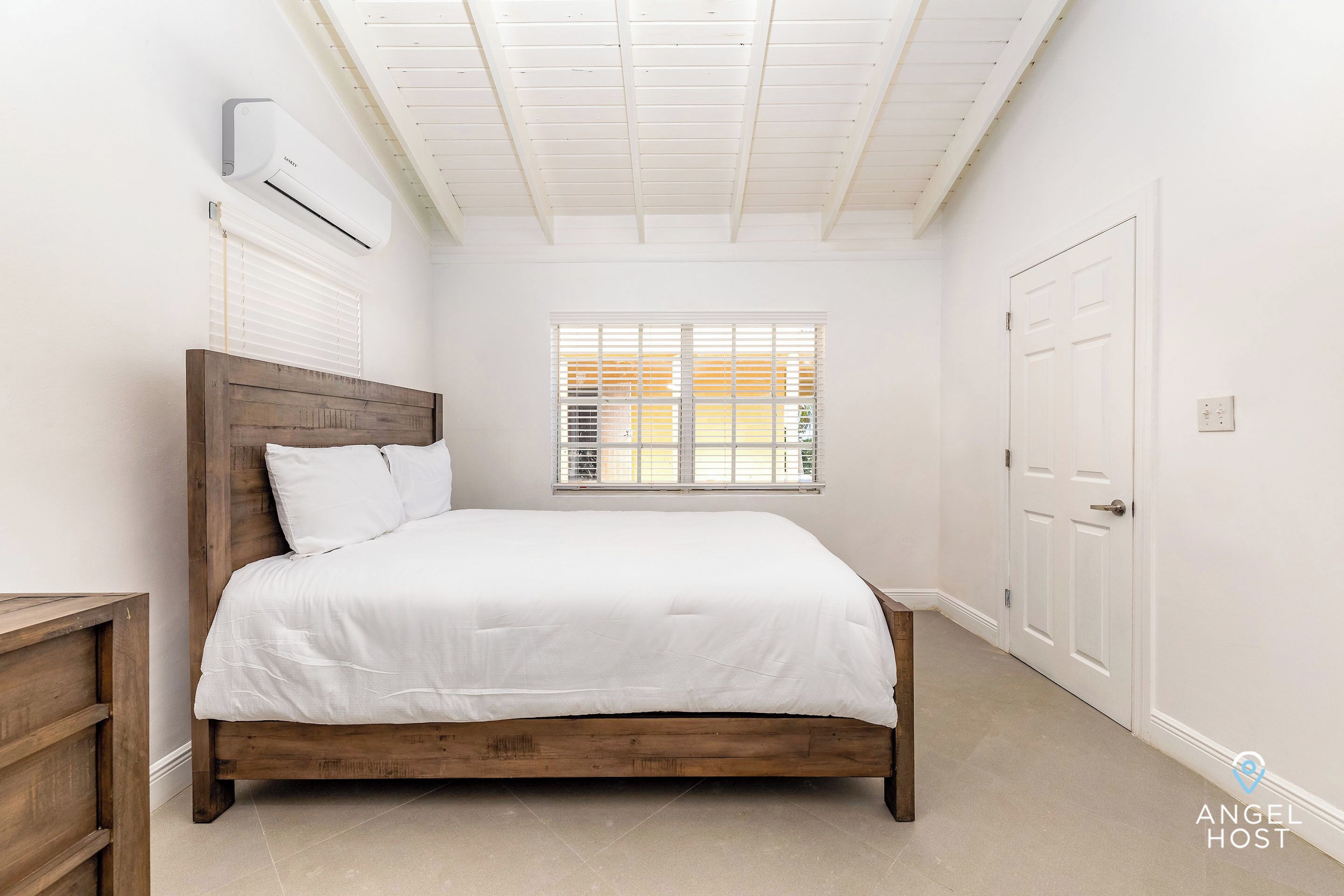 A bedroom with a wooden bed, white bedding, a window with blinds, an air conditioning unit, and a white door.