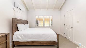 A bedroom with a wooden bed, white bedding, a window with blinds, an air conditioning unit, and a white door.