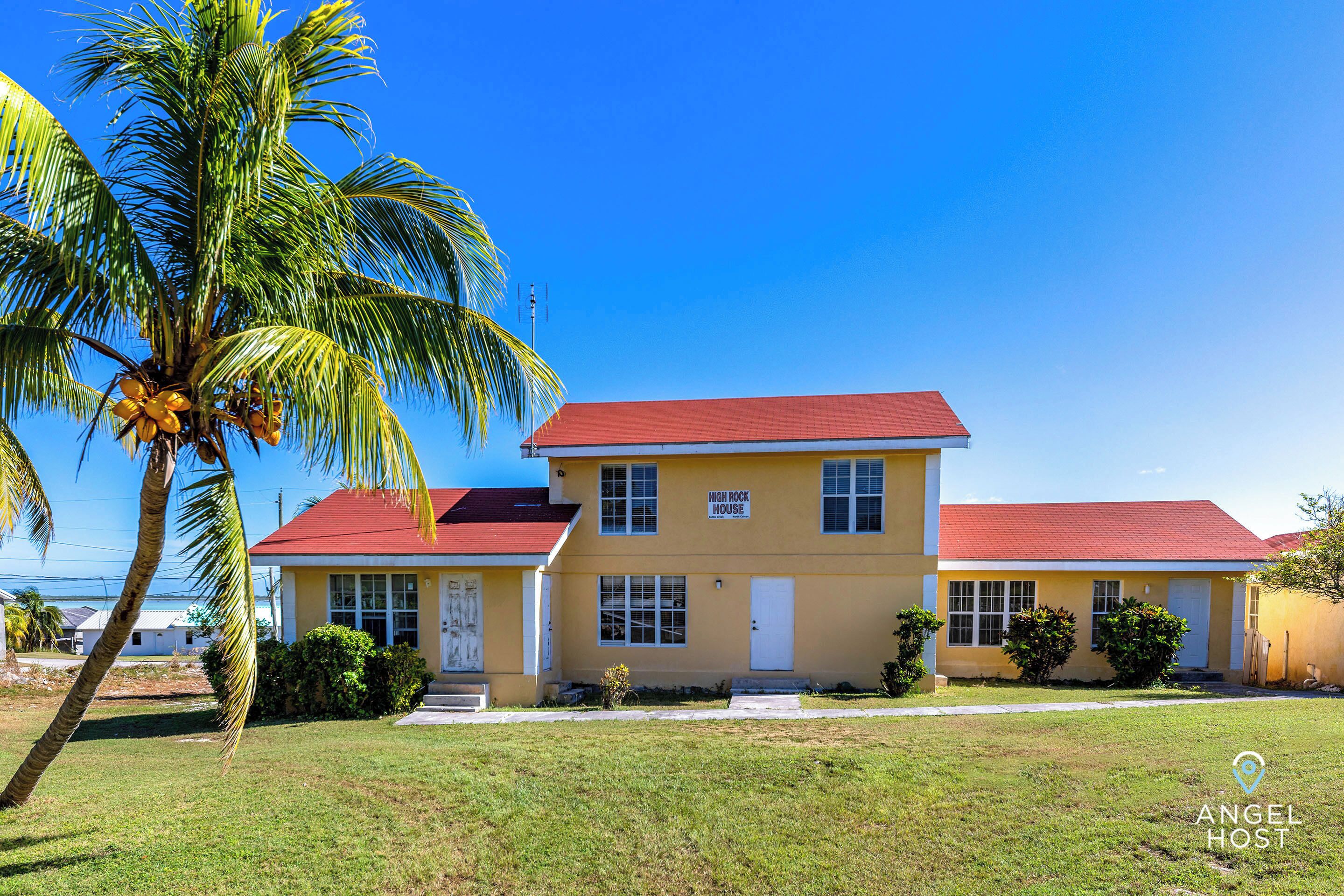 A two-story yellow building with a red roof, labeled "HIGH ROCK HOUSE," surrounded by a grassy lawn and palm trees.