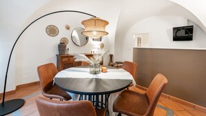 Een eetgedeelte met een ronde tafel, bruine stoelen, een geweven hanglamp en een aan de muur bevestigde televisie.