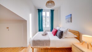A bedroom with a bed, a nightstand, a lamp, a window with curtains, and a view of buildings outside.