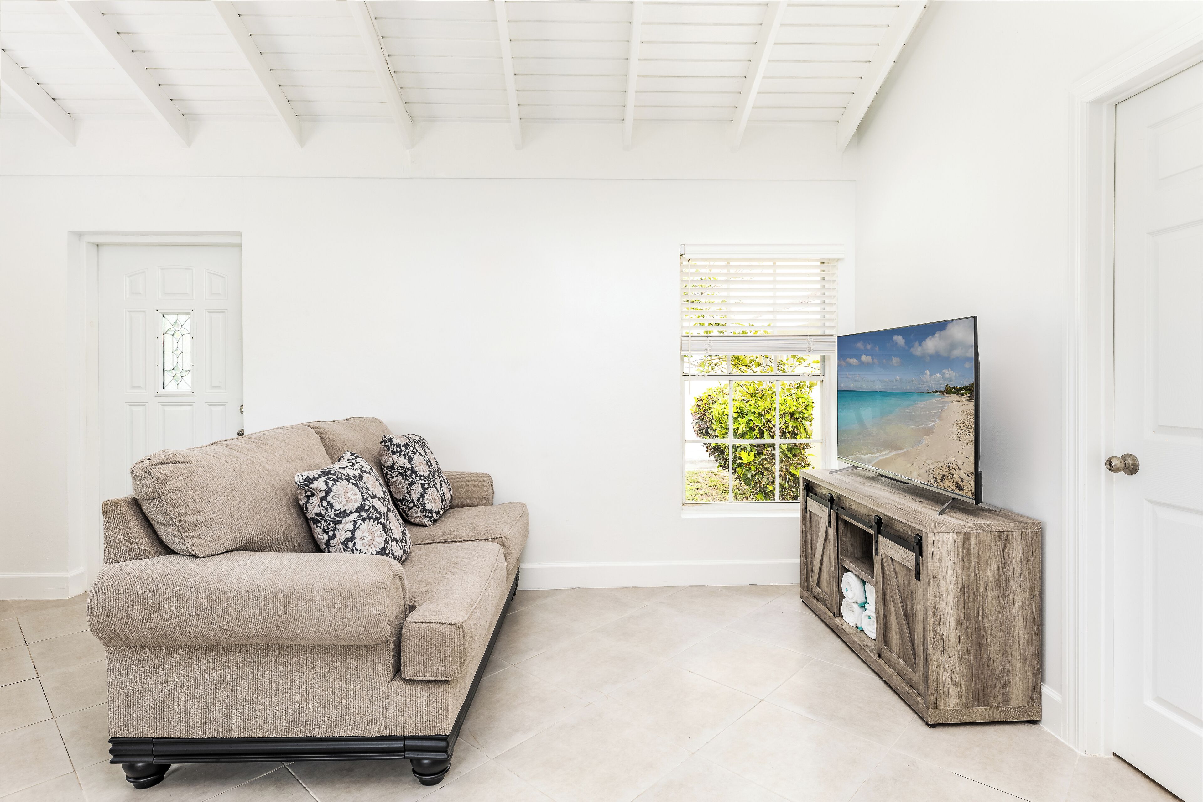 A living room with a beige sofa, a wooden TV stand, and a window with a view of greenery.