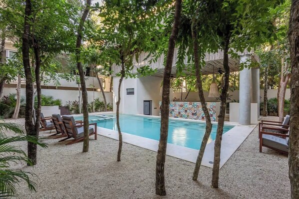A pool area with lounge chairs, trees, and a building with a tiled wall.