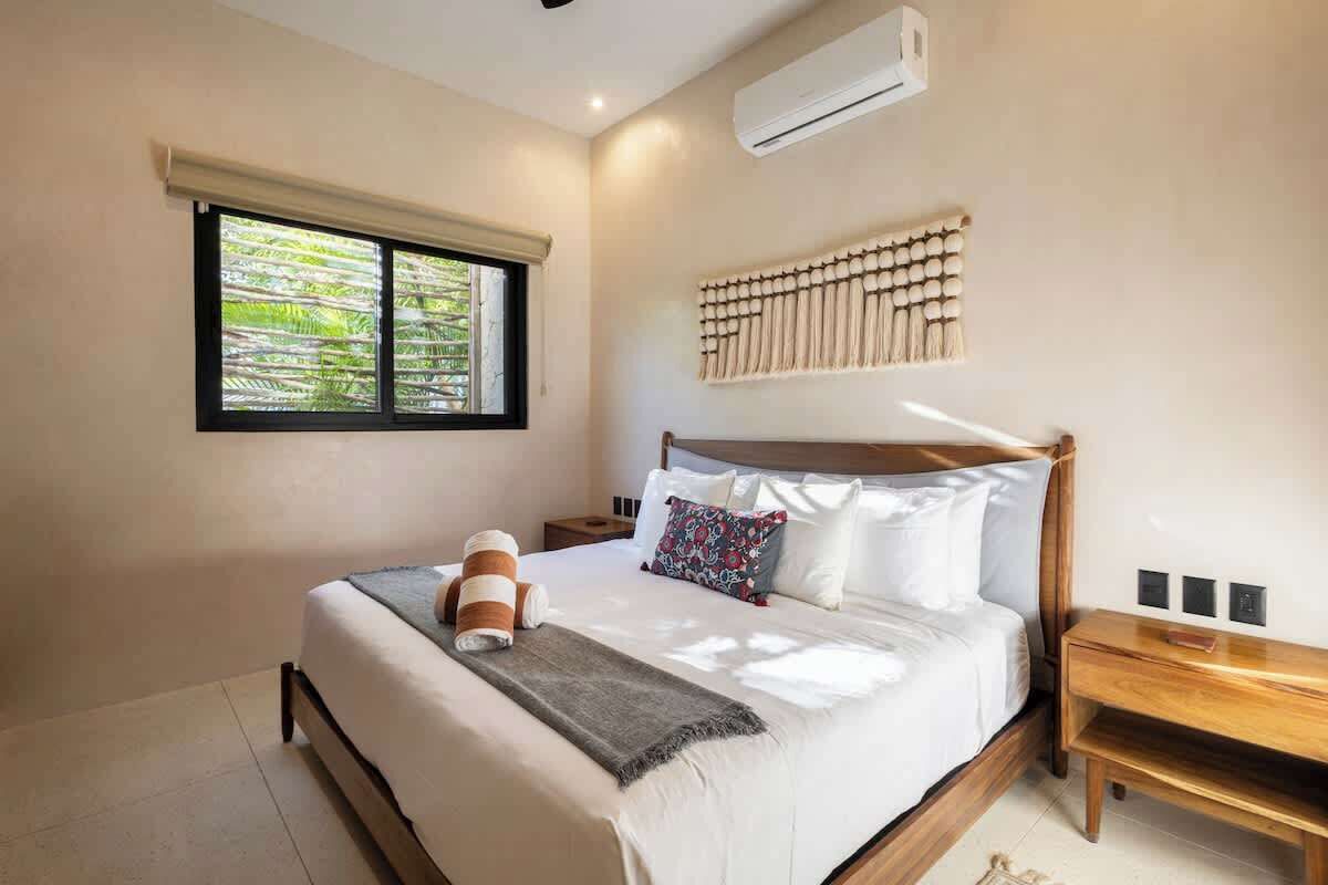 Un dormitorio con cama, ventana con persianas, aire acondicionado, mesita de noche y lámpara de pared.