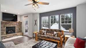 A living room with a stone fireplace, a large flat-screen TV, a sofa with patterned upholstery, and a ceiling fan. - Schweitzer Mountain 3 bd Getaway (Sandpoint)