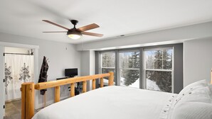 A bedroom with a wooden bed frame, a ceiling fan, and a large window with a view of trees. - Schweitzer Mountain 3 bd Getaway (Sandpoint)