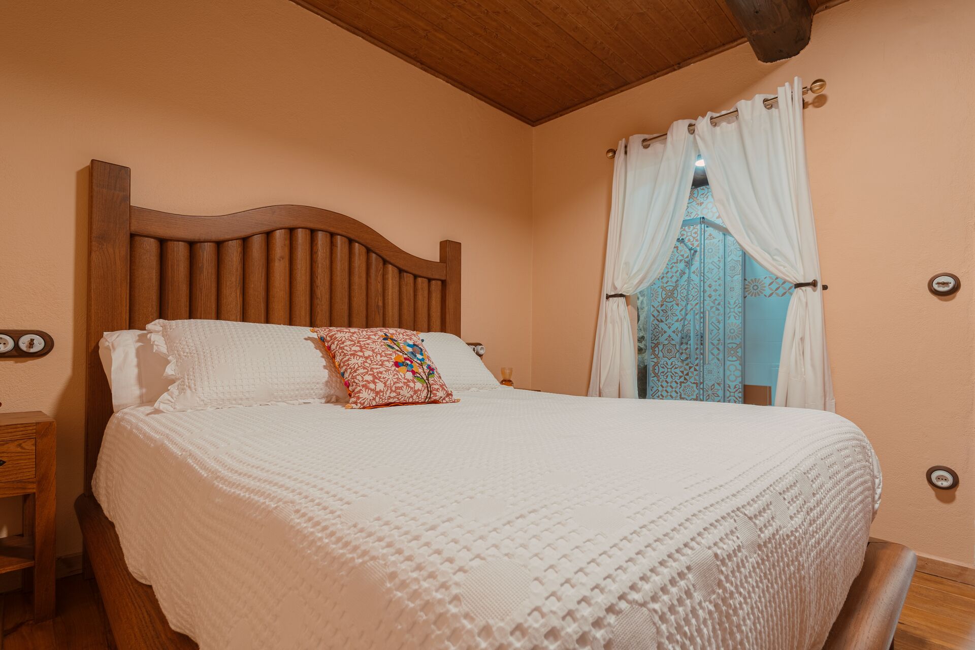 A bedroom with a wooden headboard, a bed with white bedding, a nightstand, and a window with curtains.