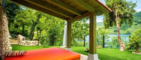Una pérgola de madera con un banco rojo y pilares de piedra, con vista a un césped verde y exuberante y un lago.