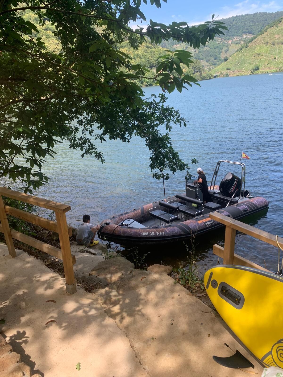 Ein Boot liegt an einem See festgemacht, eine Person ist an Bord, ein gelbes Kajak steht in der Nähe und eine Holzterrasse führt zum Wasser.