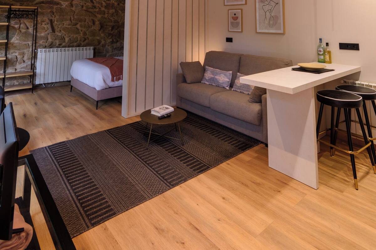 A modern living room with a stone wall, wooden beams, a grey sofa, a white coffee table, and a small bar area.