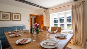 Una sala da pranzo con tavolo in legno apparecchiato per sei persone, quadri alle pareti e vista sul giardino attraverso ampie finestre.