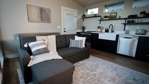 A modern living room with a dark grey sofa, a white door, and a kitchen area with a dishwasher and countertop. - Charming cabin with full RV resort access incl, WiFi in welcoming South Weber (South Weber)