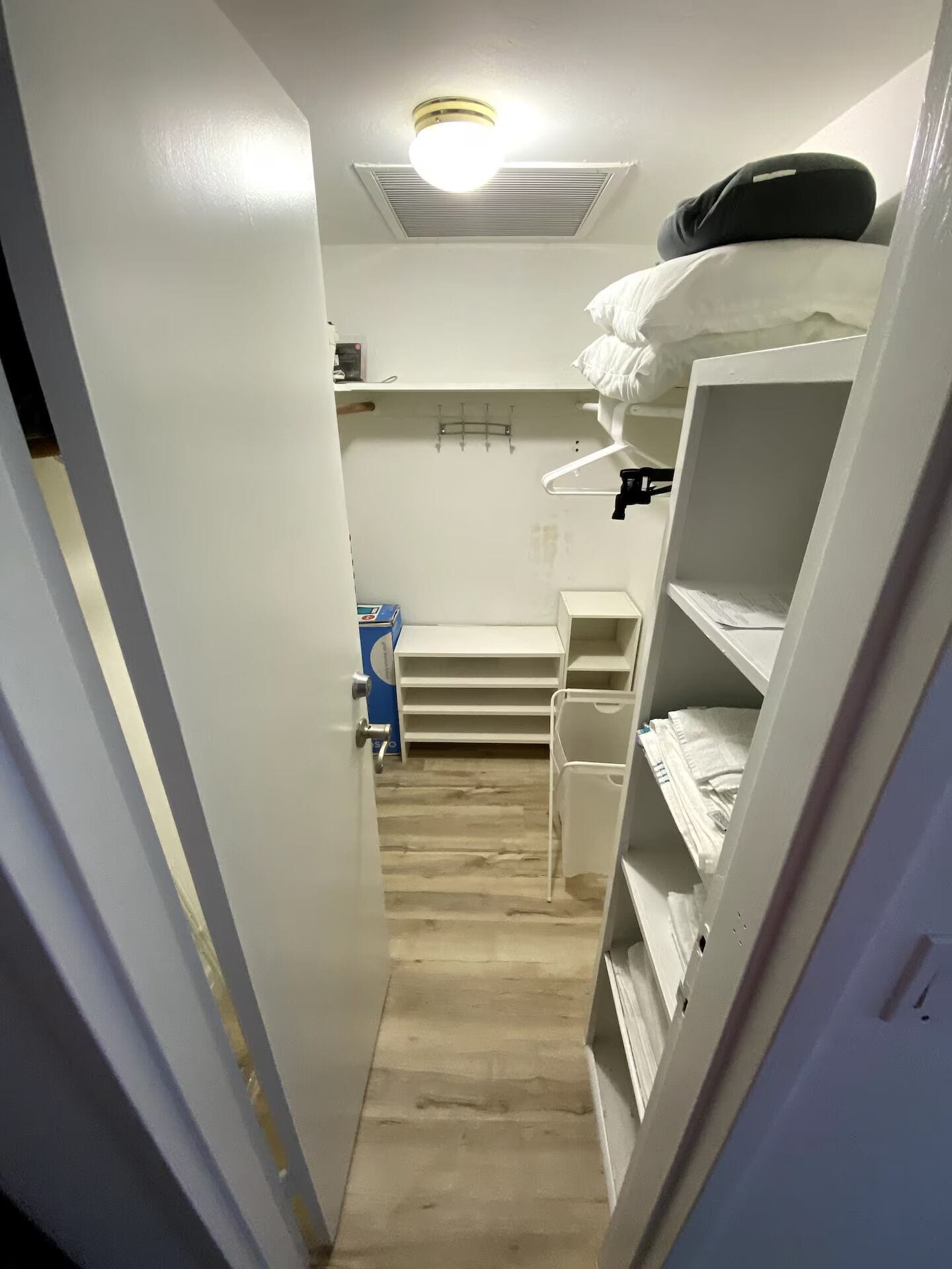 Un pasillo estrecho con paredes blancas, una luz en el techo y una puerta a la izquierda. Se ven estantes y una mesita pequeña a la derecha.