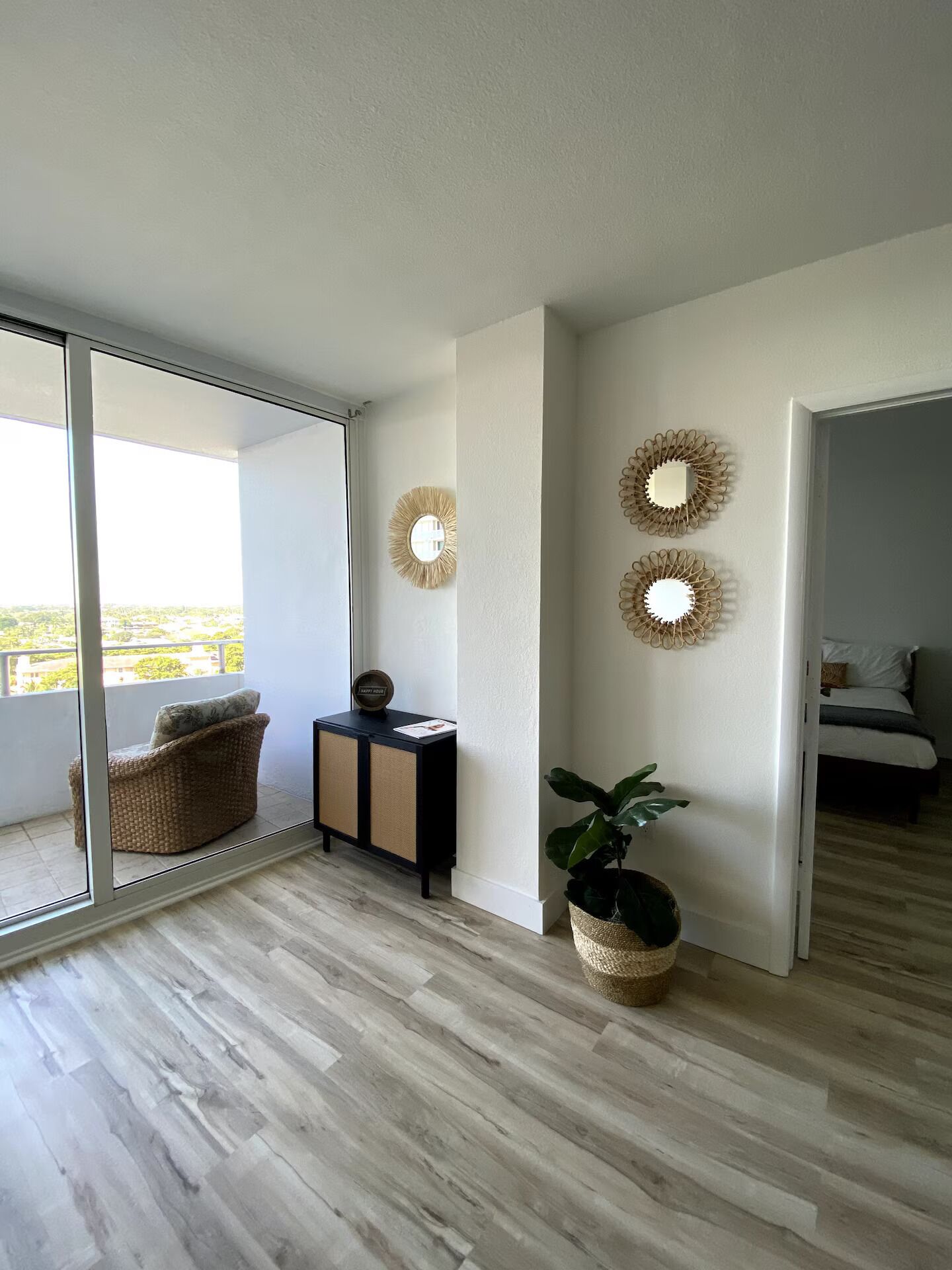 Un salon moderne doté d’une grande porte-fenêtre coulissante donnant sur un balcon, d’un fauteuil en osier, d’une petite console noire et d’une plante en pot.