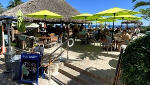 Eine Strandbar im Tiki-Stil mit Außenbestuhlung und Sonnenschirmen, einem mit Reeds gedeckten Dach und einem Schild für Bud Light.