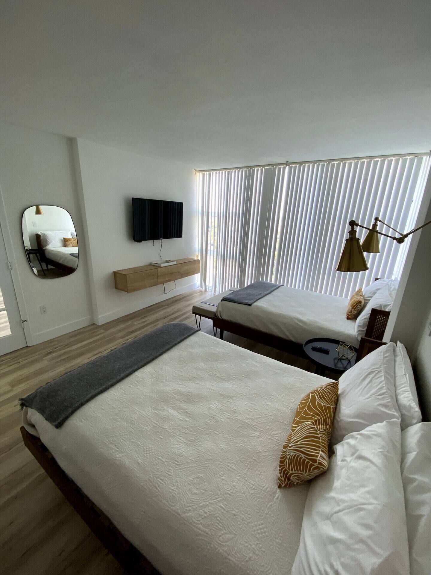 Quarto de hotel moderno com uma cama grande, mesa de cabeceira, televisão na parede e janela com persianas verticais.
