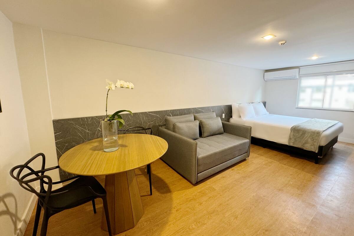 Ein modernes Zimmer mit einem runden Holztisch, einem grauen Sofa, einem Bett und einem Fenster.