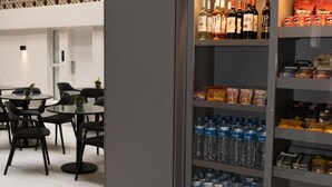Un interior moderno con un refrigerador de autoservicio repleto de agua embotellada, cerveza y vino. En primer plano, sillas negras y una mesa, y un papel tapiz estampado al fondo.