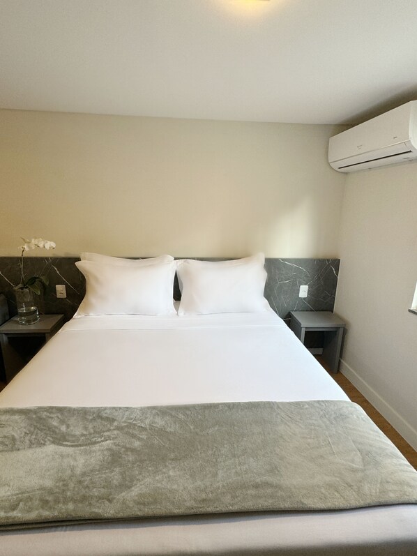 Una cama bien hecha con sábanas blancas y un plaid gris, una mesita de noche con un jarrón de flores y un cabecero con estampado de mármol.
