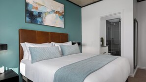 A modern bedroom with a large bed, a headboard, a nightstand, a bathroom with a glass shower, and a wall-mounted abstract painting. - Luxury Century City Apartment | Lagoon Views (Cape Town)