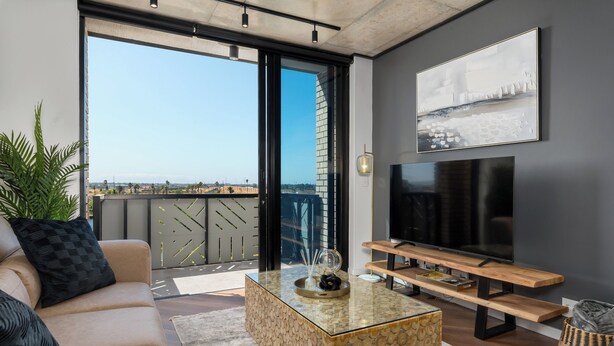 A modern living room with a sofa, coffee table, TV, and a balcony with a view. - Luxury Century City Apartment | Lagoon Views (Cape Town)