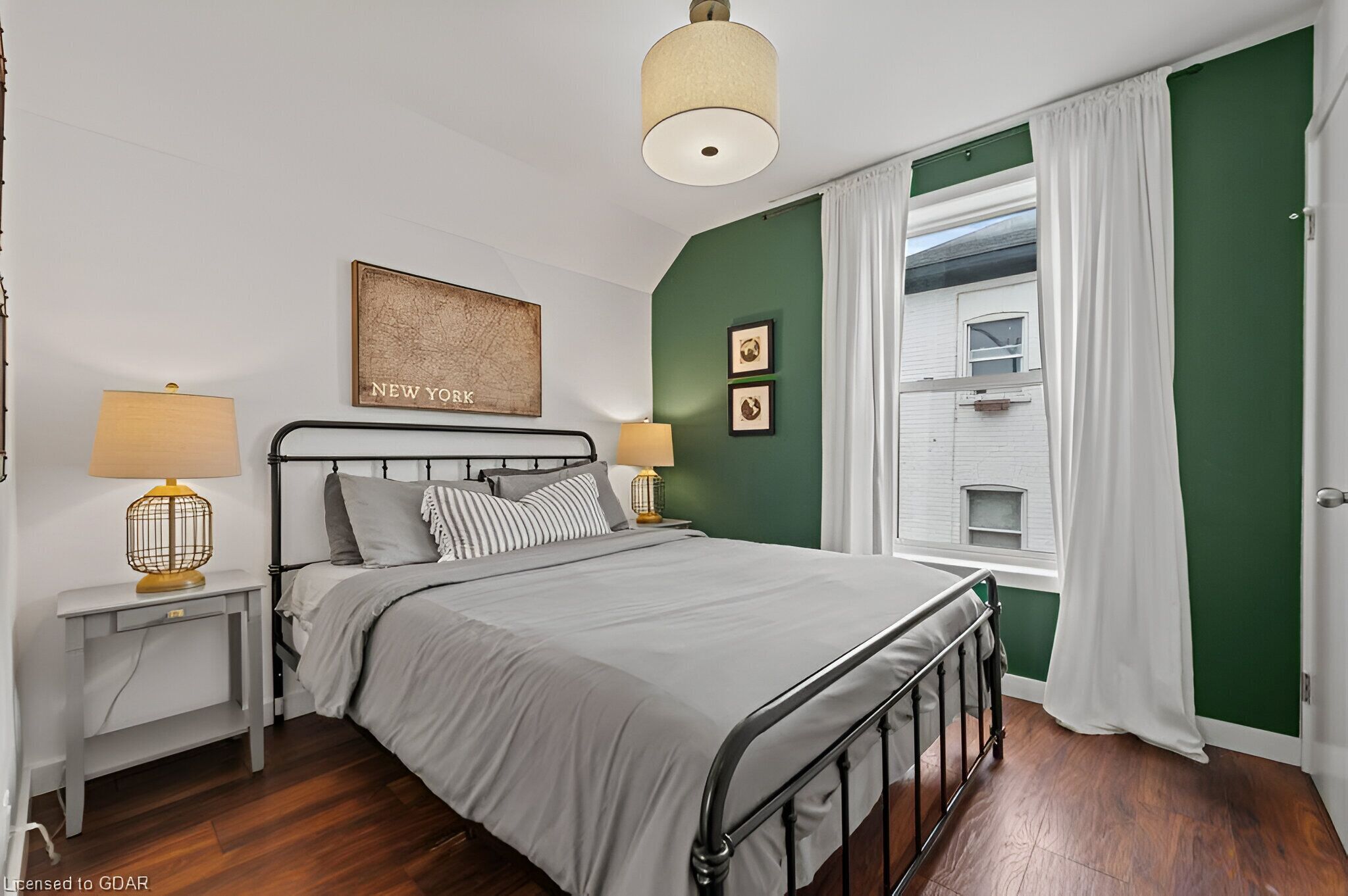 A bedroom with a bed, two bedside tables with lamps, a window with curtains, and a wall with a framed picture and two smaller pictures.