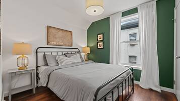 A bedroom with a bed, two bedside tables with lamps, a window with curtains, and a wall with a framed picture and two smaller pictures.