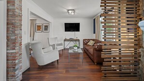 Een woonkamer met een witte bank, een bruine leren hoekbank en een salontafel. Er is een televisie aan de wand bevestigd en een houten lambrisering aan de rechterkant.