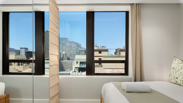 A hotel room with a bed, a wooden chair, and large windows offering a city view. - Foreshore Place | Modern Studio | Netflix (Cape Town)