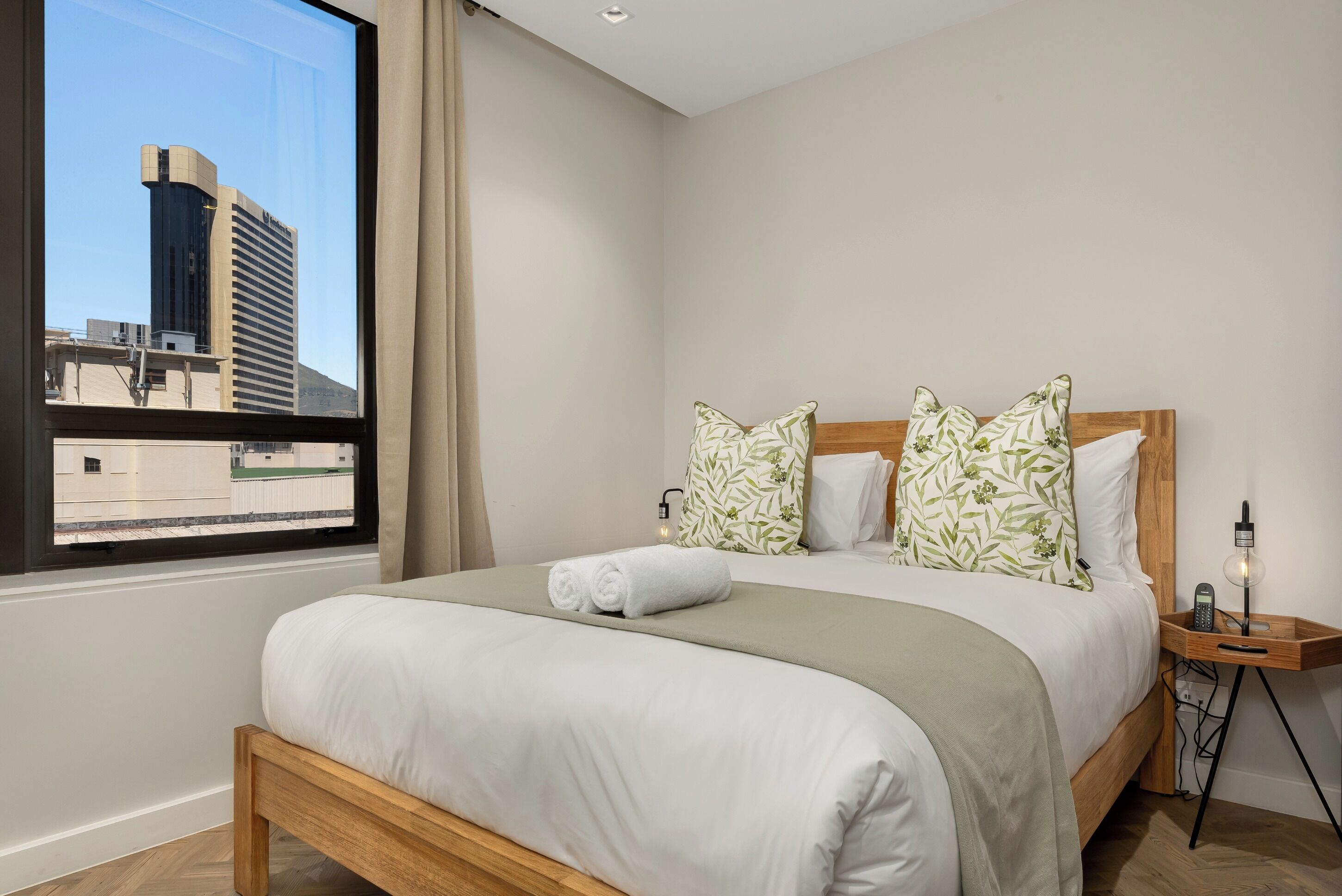 A neatly made bed with a green bedspread and white linens, a window with a view of a building, and a bedside table with a lamp.