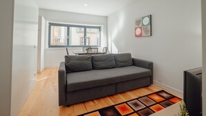 A modern living room with a grey sofa, a wooden floor, a large window, and a dining area in the background. - Downtown Sunny Apartment by Vacationy (Porto)