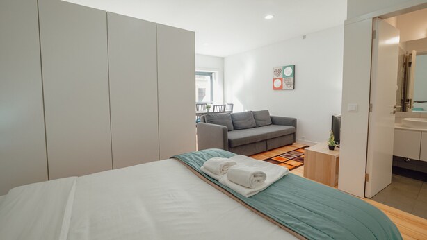 A modern hotel room with a large bed, a grey sofa, a small table with a plant, and a bathroom visible through an open door. - Downtown Sunny Apartment by Vacationy (Porto)