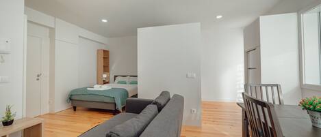 A modern, minimalist bedroom with a bed, a grey sofa, a wooden dining table, and a small plant.