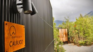 A metal structure with a sign reading "outscape CALBUCO" and a view of a natural landscape with trees and a wooden structure. - Outscape Lago Chapo (Puerto Montt)