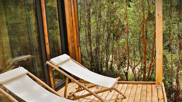 A wooden deck with two white lounge chairs facing a forest view through large windows. - Outscape Lago Chapo (Puerto Montt)