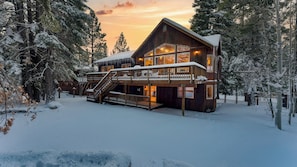 A wooden lodge with a balcony and stairs, surrounded by snow-covered trees. - Cherry Hills Spacious Tahoe Cabin With Deck, Fireplace & Open Interiors (South Lake Tahoe)