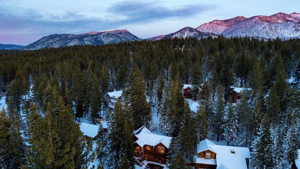 A log cabin surrounded by a dense forest with snow-covered ground and mountains in the background.