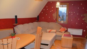 A cozy living room with a sectional sofa, a dining table with chairs, a small wooden cabinet, and a window with a view of trees.