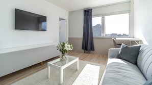 A modern living room with a flat-screen TV mounted on the wall, a glass coffee table with a vase of flowers, a grey sofa, and a large window with curtains.