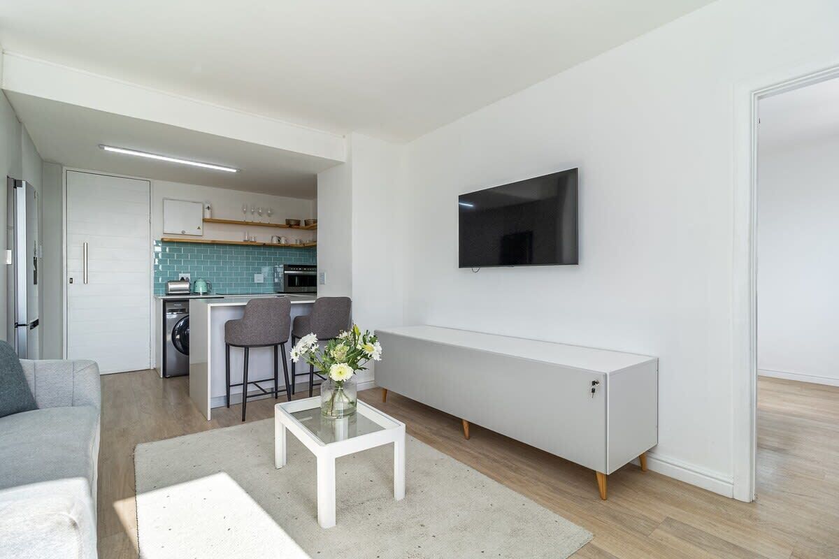 A modern living room with a sofa, a coffee table with a vase of flowers, a TV mounted on the wall, and a kitchen in the background.