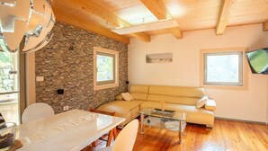 Una sala de estar moderna con pared de piedra, vigas de madera en el techo, una mesa de centro de vidrio, un sofá color beige y un televisor de pantalla plana.